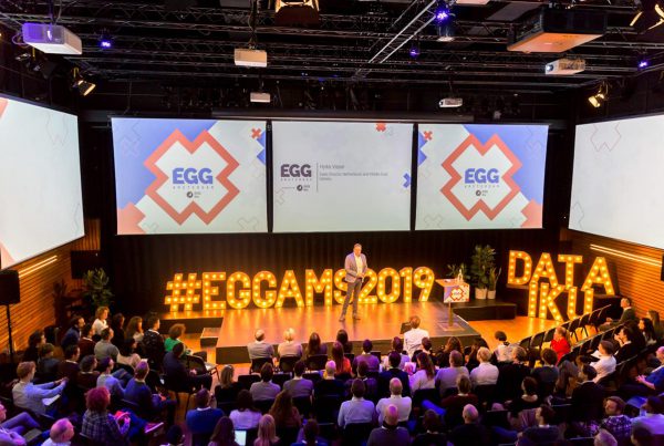 Dit is een afbeelding van de openingsfilm of kickoff van EGG Amsterdam 2019 #EGGAMS2019 dat in Pakhuis de Zwijger werd gehouden. We zien Hylke Visser het publiek aanspreken. Hij staat voor de videoschermen met daarop de huisstijl van EGG Amsterdam, bestaande uit drie Amsterdamse kruizen en blauwe of roze logo van EGG Amsterdam. Daaronder het logo in donkerblauw van Dataiku. Met mooie lampen in houten frames lezen we #EGGAMS2019. De stijl van EGG Amsterdam is volledig ontwikkeld door Activates merkversterkend reclamebureau uit Sassenheim in opdracht van Dataiku en Karin Heppener van Heppener makes it happen!